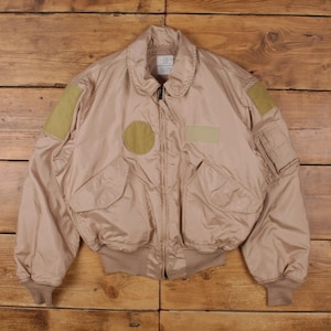 May include: A tan bomber jacket with multiple pockets, patches, and a ribbed hem and cuffs. The jacket has a zip-up front and a collared neckline. The patches are olive green and tan. The jacket is made of a smooth material.
