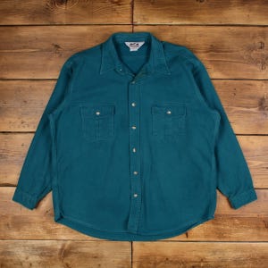 May include: Teal-colored long-sleeve shirt with two chest pockets and button-down closure. The shirt has a classic collar and is made of a durable fabric. The shirt is laid flat on a wooden surface.