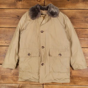 Puede incluir: Una chaqueta larga de manga larga de color beige con cuello de piel sintética marrón y múltiples cierres de botón. La chaqueta tiene dos bolsillos grandes con solapa en la parte delantera.