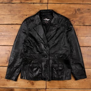May include: Black leather blazer with a notched lapel, two front pockets, and a two-button fastening. The jacket has a tailored fit and is displayed on a wooden surface. The label inside the blazer reads "mxma".