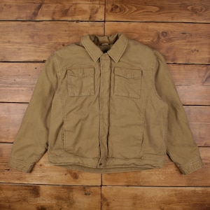 May include: A tan-colored jacket featuring a classic collar, long sleeves, and two chest pockets with flaps. The garment has a central front closure with a concealed placket and visible stitching details along the seams and hemline.