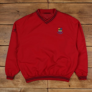 Puede incluir: Un cortavientos de golf rojo con cuello en V y mangas largas. La chaqueta tiene un ribete a rayas azul y negro en el cuello, los puños y la cintura. Un emblema bordado colorido está en el pecho izquierdo.