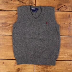 May include: A grey V-neck sweater vest with the Polo by Ralph Lauren logo. The vest features a red logo on the chest and is made of a soft material. The vest is sleeveless and has a ribbed hem.