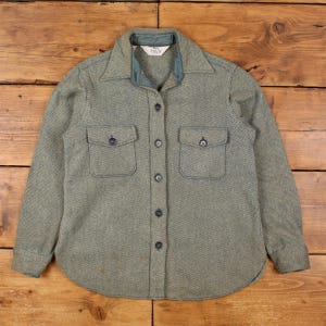 May include: A green and gray button-down shirt with two chest pockets. The shirt has a collared neckline and a rounded hem. The fabric has a textured appearance. The shirt is buttoned up and appears to be made of a thick, warm material.