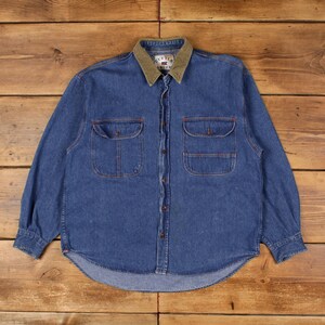 May include: A blue denim shirt with a green collar and two chest pockets. The shirt has long sleeves and a button-down front. The brand name "CUTTER & BUCK" is visible on the collar.