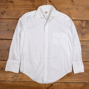 Puede incluir: Una camisa blanca de manga larga con cuello clásico y un bolsillo en el pecho. La camisa tiene puños abotonados y un dobladillo curvo. La tela parece ser de algodón o lino ligero. La camisa está tendida sobre una superficie de madera.