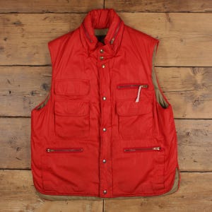 May include: A bright red sleeveless vest featuring a high collar, a front zipper closure, and a snap-button placket. The garment includes two chest pockets with flaps, two lower zippered pockets, and a tan lining visible along the inner edges.