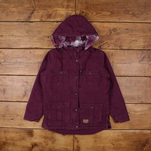 May include: A burgundy hooded jacket with a plaid-lined hood. The jacket features four front pockets, a zipper closure, and button details. The jacket has a small patch on the lower right side.