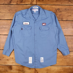 May include: A light blue long-sleeve button-down work shirt featuring a pointed collar, two chest pockets, and a central button placket. The shirt displays a name patch reading Jason, a logo patch reading Wt, and two white rectangular labels at the hem.