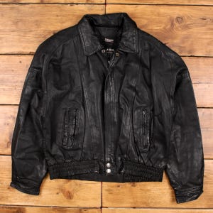 Puede incluir: Chaqueta de cuero negra con cuello, dos bolsillos delanteros y cierre de cremallera. La chaqueta tiene dobladillo y puños acanalados. La chaqueta se muestra sobre una superficie de madera.