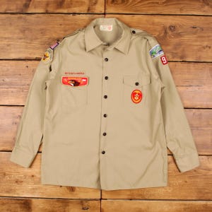 May include: A tan long-sleeve button-down shirt featuring Boy Scouts of America patches, including an eagle emblem on the right chest pocket, a circular red and yellow badge on the left pocket, and various shoulder patches on both sleeves.