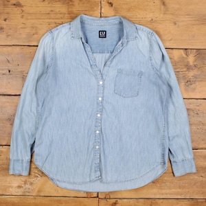 May include: Pale blue denim shirt with long sleeves, a button-down front, and a single chest pocket. The shirt has a classic collar and the "GAP DENIM" logo tag. The shirt is laid flat on a wooden surface.