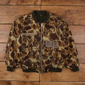 Puede incluir: Chaqueta bomber de camuflaje vintage con cierre de cremallera completo. La chaqueta presenta un patrón de formas marrones y tostadas sobre un fondo verde claro. Tiene dos bolsillos delanteros y cuello y puños verde oscuro.