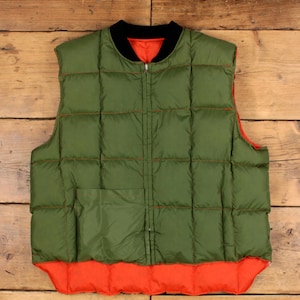 May include: A quilted olive green puffer vest featuring a black ribbed collar, a full-length front zip closure, and a square patch pocket on the lower left side. The interior lining is a bright orange colour, visible at the hem and collar.
