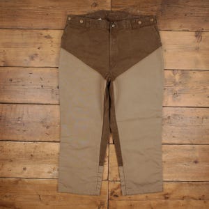 May include: Brown and tan pants featuring a V-shaped color-blocked design. The upper portion is dark brown, while the lower leg sections are a lighter tan color. The pants include a button-front waistband with belt loops and side pockets.