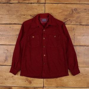 Puede incluir: Una camisa burdeos de manga larga con dos bolsillos en el pecho y cuello. La camisa tiene botones frontales y botones dorados. La tela parece ser un material suave y tejido.