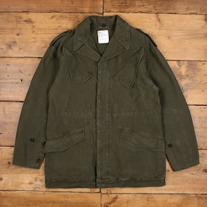 Puede incluir: Chaqueta de estilo militar verde oliva con cierre de botones, dos bolsillos en el pecho y dos bolsillos inferiores. La chaqueta tiene cuello y charreteras. Una pequeña etiqueta es visible cerca del cuello. La chaqueta es de tela resistente.