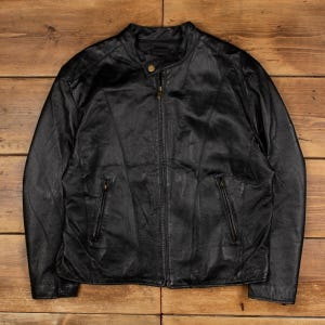 Puede incluir: Chaqueta de cuero negro con cuello alzado y cierre de cremallera frontal. La chaqueta tiene dos bolsillos con cremallera y un cierre de botón en el cuello. El cuero parece ligeramente desgastado, dándole un aspecto vintage.