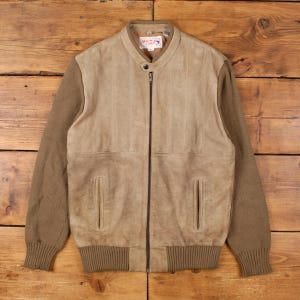 May include: A tan suede jacket with brown knit sleeves and a ribbed hem and cuffs. The jacket has a zip-up front, two front pockets, and a small collar. The jacket is laid flat on a wooden surface.