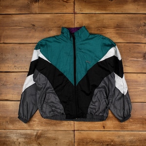 Könnte beinhalten: Eine Vintage-Jacke mit Reißverschluss in Teal, Schwarz, Weiß und Grau. Die Jacke hat einen hohen Kragen mit lila Futter und ein kleines Logo auf der Brust. Die Jacke ist aus einem leichten Material, möglicherweise Nylon.