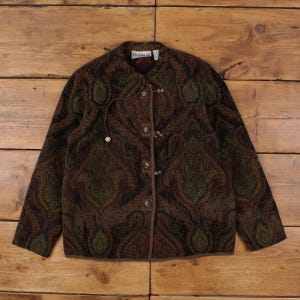 May include: A brown paisley print jacket with a round collar and toggle closures. The jacket features a detailed pattern in shades of brown, green, and red. The jacket is displayed on a wooden surface.