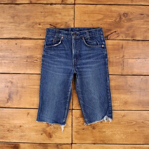 May include: Blue denim shorts with a button fly and frayed edges. The shorts have classic five-pocket styling and a worn appearance. The garment is displayed on a wooden surface.