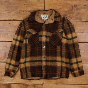 May include: A long-sleeve button-up shirt featuring a brown, tan, and dark green plaid pattern. The shirt has a tan collar, two chest pockets with buttoned flaps, and a Woolrich brand label sewn onto the inside of the collar area.