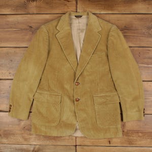 Könnte beinhalten: Ein beigefarbener Cord-Blazer mit steigendem Revers, zwei braunen Knöpfen in der vorderen Mitte und zwei großen aufgesetzten Taschen an der Taille. Die Ärmel sind mit drei kleinen braunen Knöpfen an den Manschetten versehen und innen beige gefüttert.