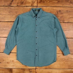 May include: A long-sleeve button-down shirt in a solid teal colour. The shirt features a pointed collar, a single chest pocket on the left side, and dark buttons running down the centre placket. The fabric has a soft, brushed texture.