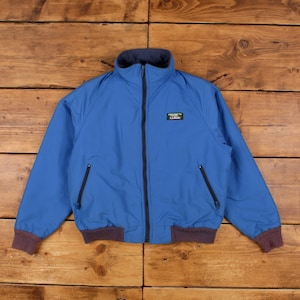 May include: A blue zip-up jacket featuring a high collar, dark blue trim, and a dark brown ribbed hem and cuffs. The jacket includes two zippered side pockets and a small rectangular L.L.Bean logo patch on the upper left chest area.