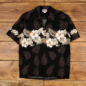 May include: A black short-sleeve button-down shirt featuring a horizontal band of white hibiscus flowers and light-colored tropical leaves across the chest. The rest of the shirt is patterned with dark brown fern-like leaf silhouettes.