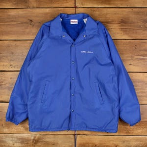 Puede incluir: Chaqueta de entrenador azul real con cierre de botones a presión, dos bolsillos y un logotipo blanco que dice "LOEB & LOEB LLP". La chaqueta tiene un forro polar blanco y azul y un cuello. La chaqueta está tendida sobre una superficie de madera.