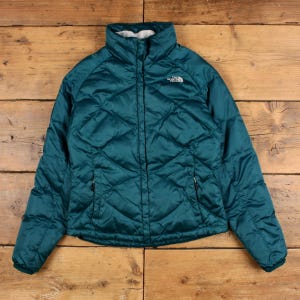 May include: A teal puffer jacket with a diamond-quilted pattern, a high collar, and a front zip closure. The North Face logo is embroidered in white on the upper left chest. The jacket features long sleeves and two side pockets with zip pulls.