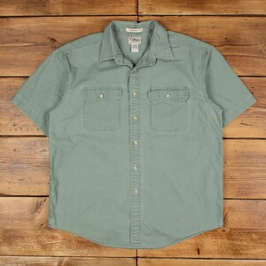 Op de afbeelding: Een korte mouwen, saliegroen button-down shirt met twee borstzakken. Het shirt heeft een kraag en is gemaakt van een duurzame stof. Het shirt ligt plat op een houten oppervlak.