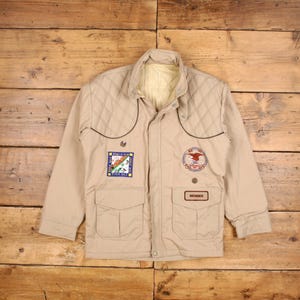 May include: Tan jacket with a quilted shoulder design, two front pockets, and a button closure. Features patches with text and a bird emblem. The jacket has a "MEMBER" tag.