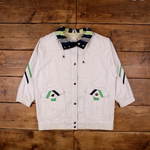 May include: White jacket with a navy, green, and yellow striped collar and pocket accents. The jacket has a button-up front, two front pockets, and green and navy stripes on the sleeves. The jacket is on a wooden surface.