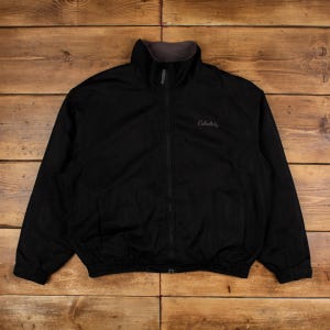 Puede incluir: Chaqueta negra con cremallera completa y cuello alto. La chaqueta tiene un cuello interior gris y el nombre de la marca "Cabela's" bordado en el pecho. La chaqueta está hecha de un material liso, posiblemente resistente al agua.