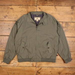 May include: A sage green zip-up jacket featuring a classic collar, long sleeves with ribbed cuffs, and two front pockets with button closures. The interior is lined with a navy, red, and white plaid fabric, and a Woolrich brand label is visible.