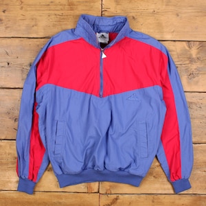 Könnte beinhalten: Eine blaue und rote Adidas Vintage-Trainingsjacke. Die Jacke hat einen halben Reißverschluss, einen Stehkragen und zwei Vordertaschen. Die obere Brust und die Ärmel sind rot, während der untere Teil blau ist. Das Adidas-Logo ist sichtbar.