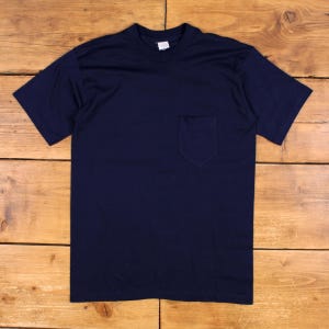 Może przedstawiać: Gładki, granatowy t-shirt z krótkim rękawem, okrągłym dekoltem i pojedynczą naszywaną kieszenią po lewej stronie klatki piersiowej. Odzież ma standardowy krój, krótkie rękawy i proste wykończenie dołu, prezentując minimalistyczny wygląd.