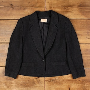 May include: A dark grey blazer with a single-button fastening and two front pockets. The jacket features a notched lapel and a tailored fit. The interior lining is black. The blazer is displayed on a wooden surface.
