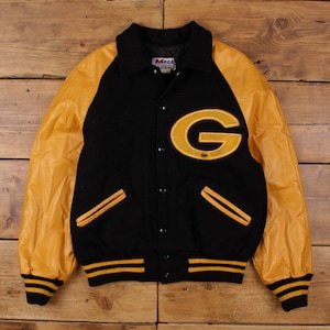 Könnte beinhalten: Schwarz-goldene Collegejacke mit großem goldenem "G"-Aufnäher. Die Jacke hat goldene Lederärmel, einen schwarzen Körper und gold-schwarz gestreifte Bündchen und Bund. Die Jacke hat eine Knopfleiste.