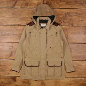 May include: A tan jacket with a hood, multiple pockets, and brown leather shoulder patches. The jacket features a full zip and a drawstring hood. Brown trim accents the pockets and cuffs. The brand name "Orvis" is visible on the chest.