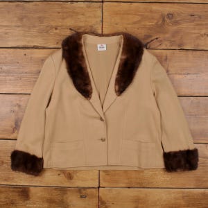 Könnte beinhalten: Beige Blazer mit braunem Pelzkragen und Manschetten. Der Blazer hat einen Einzelknopfverschluss und zwei Vordertaschen. Das Kleidungsstück wird auf einer Holzoberfläche präsentiert.