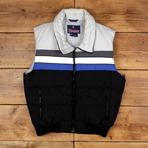 Puede incluir: Un chaleco acolchado negro, azul, gris y blanco con cremallera completa. El chaleco tiene un cuello gris y un dobladillo inferior negro. El chaleco tiene dos bolsillos negros con cremallera. La marca "Hermann's" es visible en el cuello.