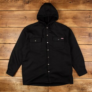 Peut inclure: Une chemise-veste à capuche noire à manches longues avec fermeture à boutons-pression, deux poches poitrine à rabat boutonné et une capuche à cordon de serrage. Un petit écusson rectangulaire est visible sur la poche poitrine droite.