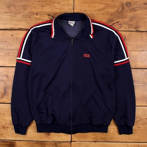 Könnte beinhalten: Marineblaue Trainingsjacke mit Reißverschluss und rot-weißen Streifen auf Schultern und Ärmeln. Das Wort "USA" ist rot auf die Brust gestickt. Mit zwei Vordertaschen.