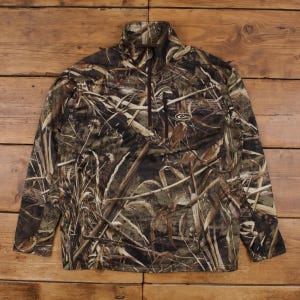May include: A long-sleeve pullover jacket featuring a camouflage pattern of brown, tan, and dark green marsh grass. The garment has a high collar, a partial front zipper, and a single zippered chest pocket with a small logo embroidered in white.