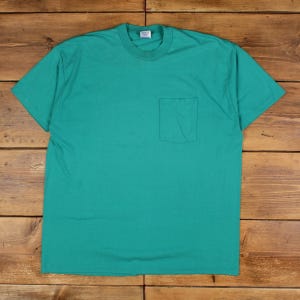 Könnte beinhalten: Ein türkisfarbenes T-Shirt mit kurzen Ärmeln und einer Brusttasche. Das Shirt hat einen Rundhalsausschnitt und ist aus einem schlichten Stoff gefertigt. Das T-Shirt liegt flach auf einer Holzoberfläche.