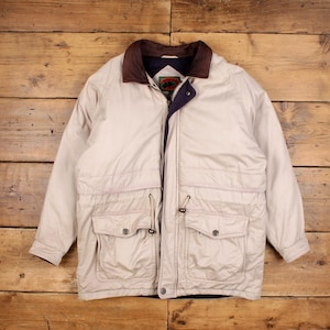 May include: A beige jacket with a brown collar and multiple pockets. The jacket has a drawstring at the waist and a dark blue lining. The jacket is laid flat on a wooden surface.